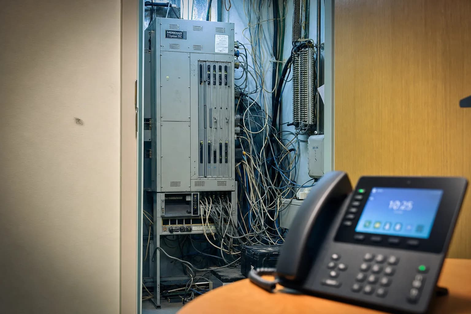 An old PBX phone system cabinet in a wiring closet next to a simple modern desk phone, comparing a pbx system vs voip